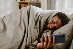 Ein Mann liegt im dunklen Bett und starrt auf sein leuchtendes Smartphone als Visualisierung von Blaulicht-Stress und fehlender Erholung.